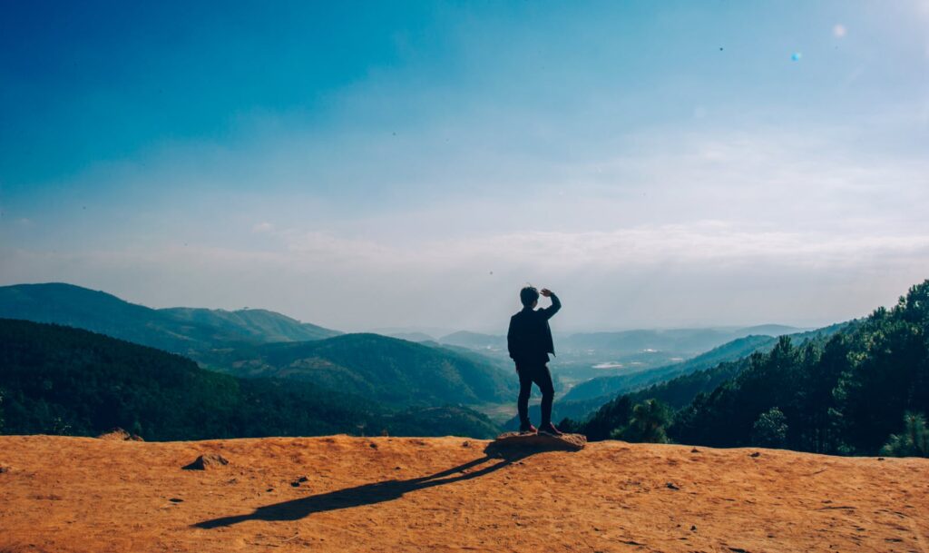 behavioral health, mental health, therapy A lone traveler stands on a hilltop, gazing over a vast mountain landscape under a clear blue sky.