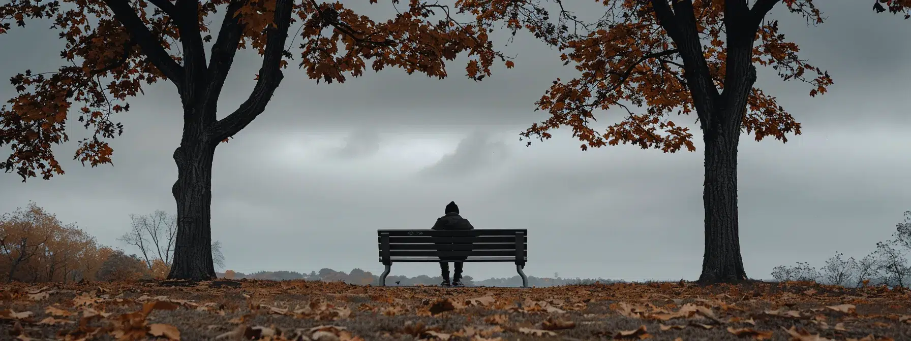 a hauntingly beautiful scene captures a solitary figure seated on a park bench under a muted gray sky, surrounded by withered autumn leaves, embodying the weight of anxiety and depression through their distant gaze and a palpable sense of introspection.