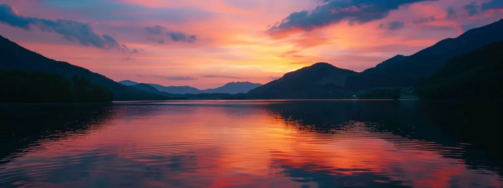 a vibrant sunset casts brilliant hues of orange and pink across a tranquil lake, with silhouetted mountains framing the horizon and lush greenery reflecting on the water's surface.