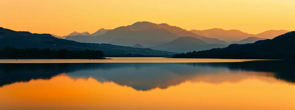 a vibrant sunset casts golden hues over a tranquil lake, reflecting the majestic silhouettes of distant mountains against a clear, serene sky.